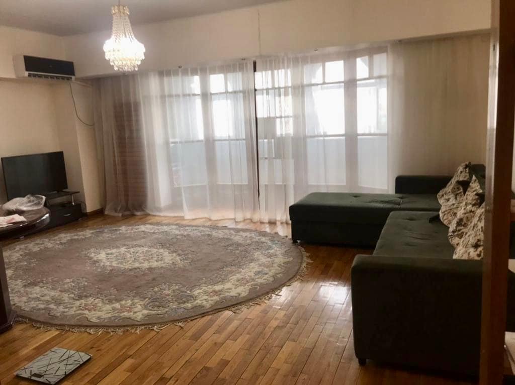 A spacious living area is designed with oak parquet flooring, featuring a large round area rug and a cozy canapé for two. Natural light filters through sheer curtains covering multiple windows, illuminating the room. A flat-screen TV is positioned on a decorative stand.