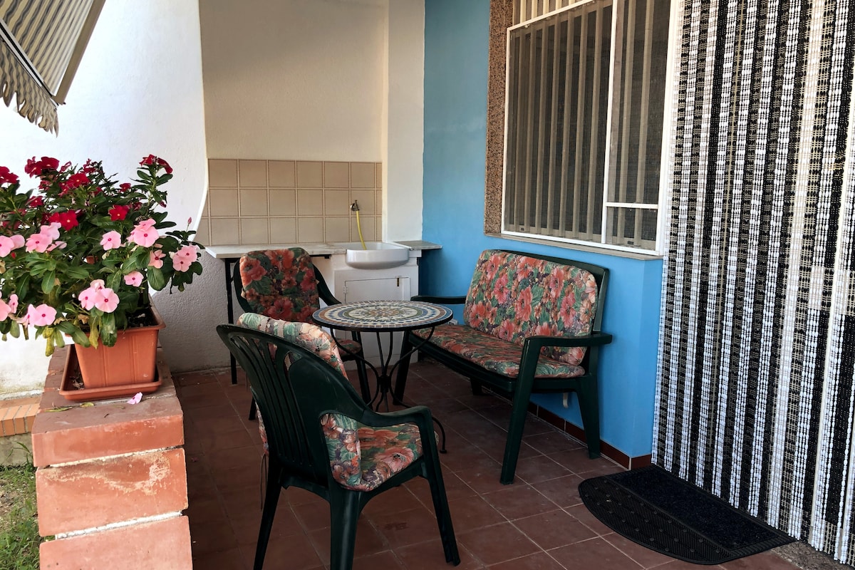 A shaded outdoor terrace features a round table surrounded by four chairs with floral cushions. A small sink is visible against the wall, and a planter with pink flowers adds a touch of color. The floor is tiled, enhancing the cozy setting.