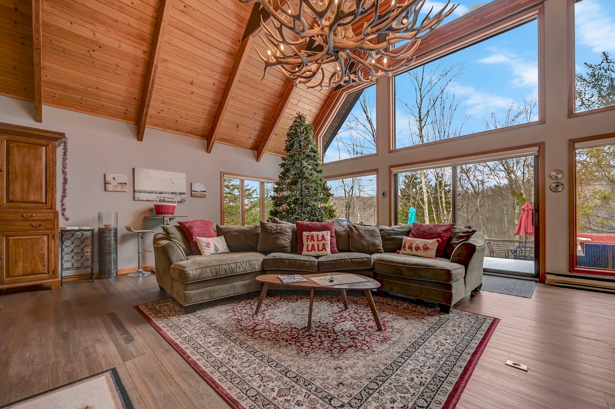 A spacious living area features high vaulted ceilings and large windows offering natural light and views of the outdoors. A comfortable sectional sofa is placed on a patterned rug, while a decorative antler chandelier hangs above. A Christmas tree is positioned nearby, enhancing the inviting setting.