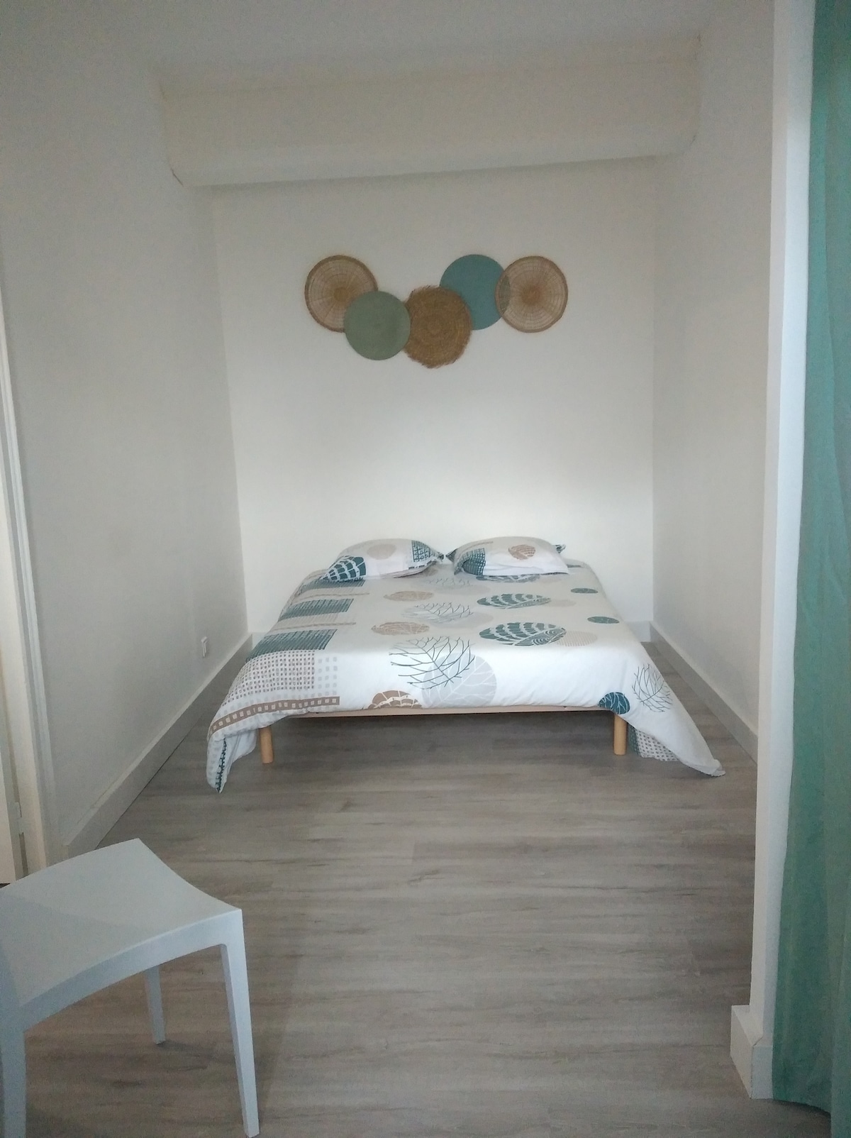 A minimalist bedroom features a spacious bed dressed in a patterned duvet. Decorative wall art composed of woven discs adds visual interest. A small chair is positioned nearby, and neutral-colored walls contribute to the room's bright, airy ambiance.