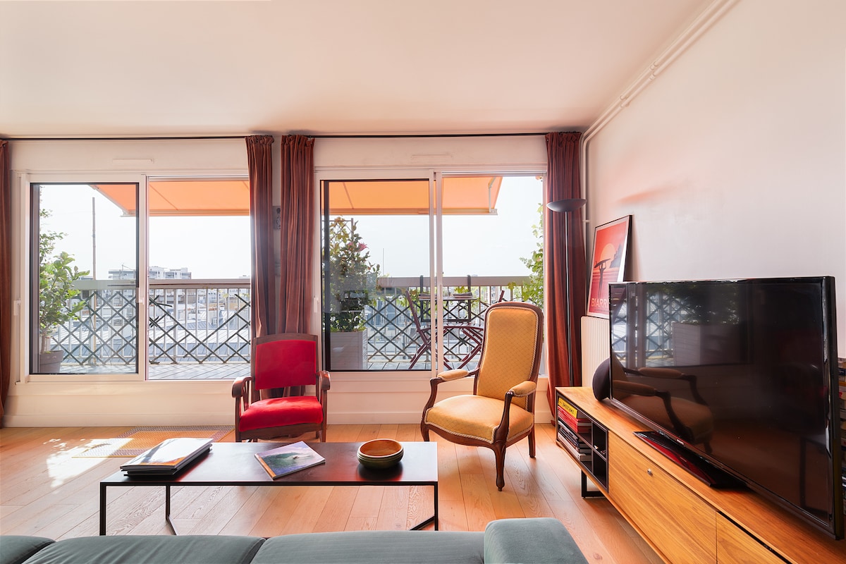 A living area features a green sofa and a wooden coffee table. Two large windows provide natural light, with vibrant orange awnings partially extended. A red armchair and a yellow armchair are included. A television sits atop a wooden media console, while plants are visible outside.