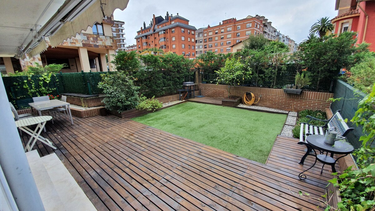 A spacious patio area features a well-maintained lawn and wooden decking. Lush greenery surrounds the space, providing privacy. A small seating area with a table and chairs, along with a barbecue grill, offers an inviting spot for relaxation or dining outdoors.