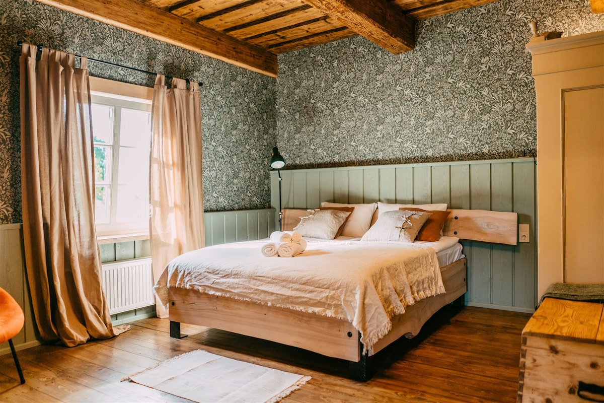 A cozy bedroom features a spacious bed with light bedding and neatly arranged towels. The walls are adorned with floral wallpaper, complemented by wooden beams and flooring. Natural light filters through a nearby window, framed by light curtains, creating a warm ambiance.