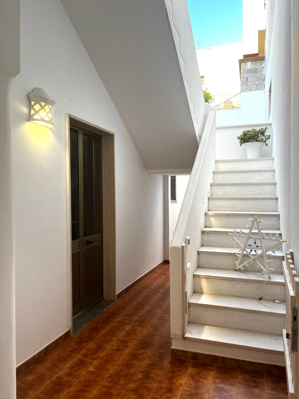A bright corridor showcases a staircase with white steps leading to an upper level. The walls are painted in a light color, complemented by a warm light fixture. A small potted plant adds a touch of greenery to the space.