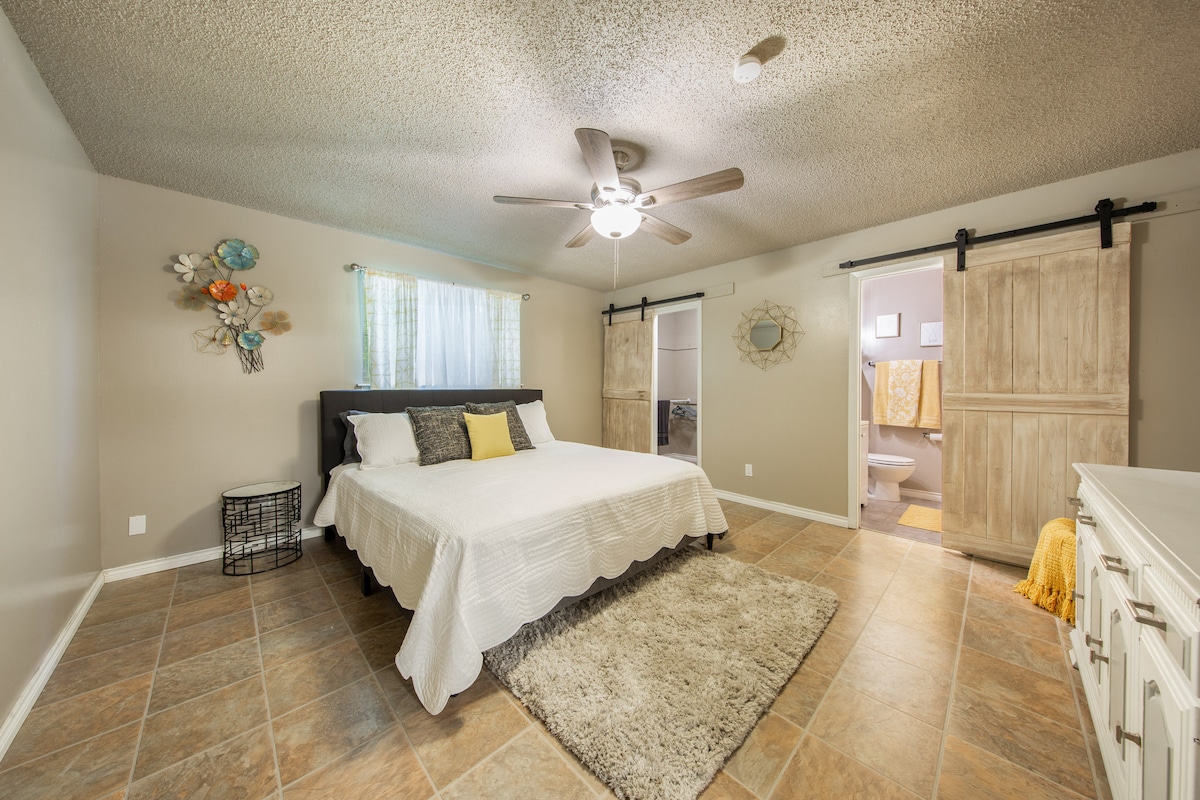A spacious bedroom features a large bed with a light-colored duvet and decorative pillows. A ceiling fan provides airflow, while soft natural light illuminates the room. Barn-style doors lead to a bathroom area, enhancing the cozy, modern design.