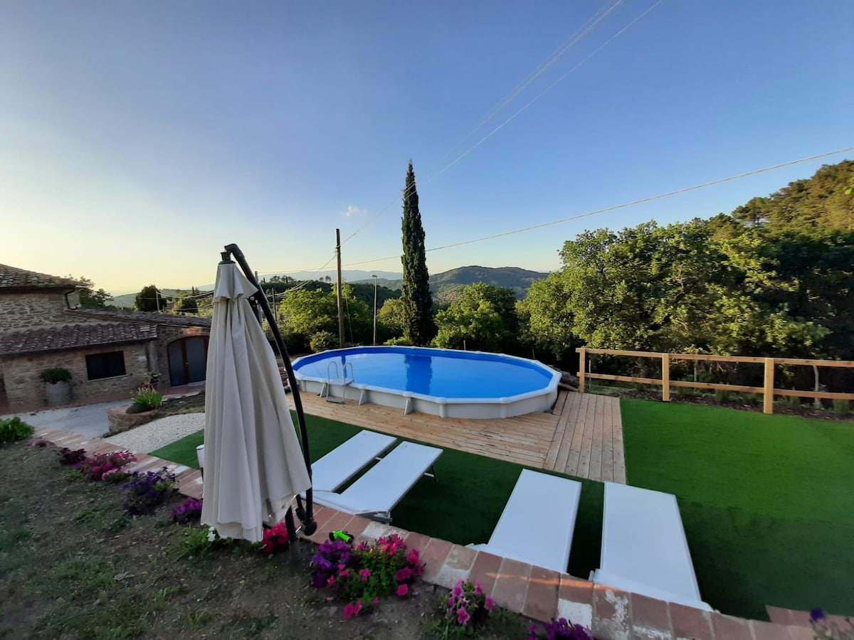 A circular pool is situated on a wooden deck, surrounded by lush greenery and colorful flowers. Sun loungers are arranged nearby, and a canopy stands to the side, providing shade. The view extends over the landscape, with rolling hills visible in the background.