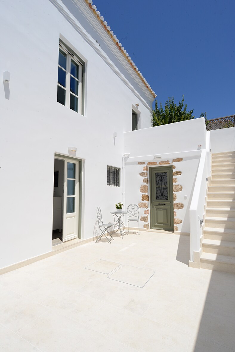 A bright outdoor area is framed by white walls, featuring a small table with two chairs. A tranquil staircase leads to an upper level, while greenery peeks above, adding a touch of nature to the scene. The sky is clear and blue, contributing to a serene setting.