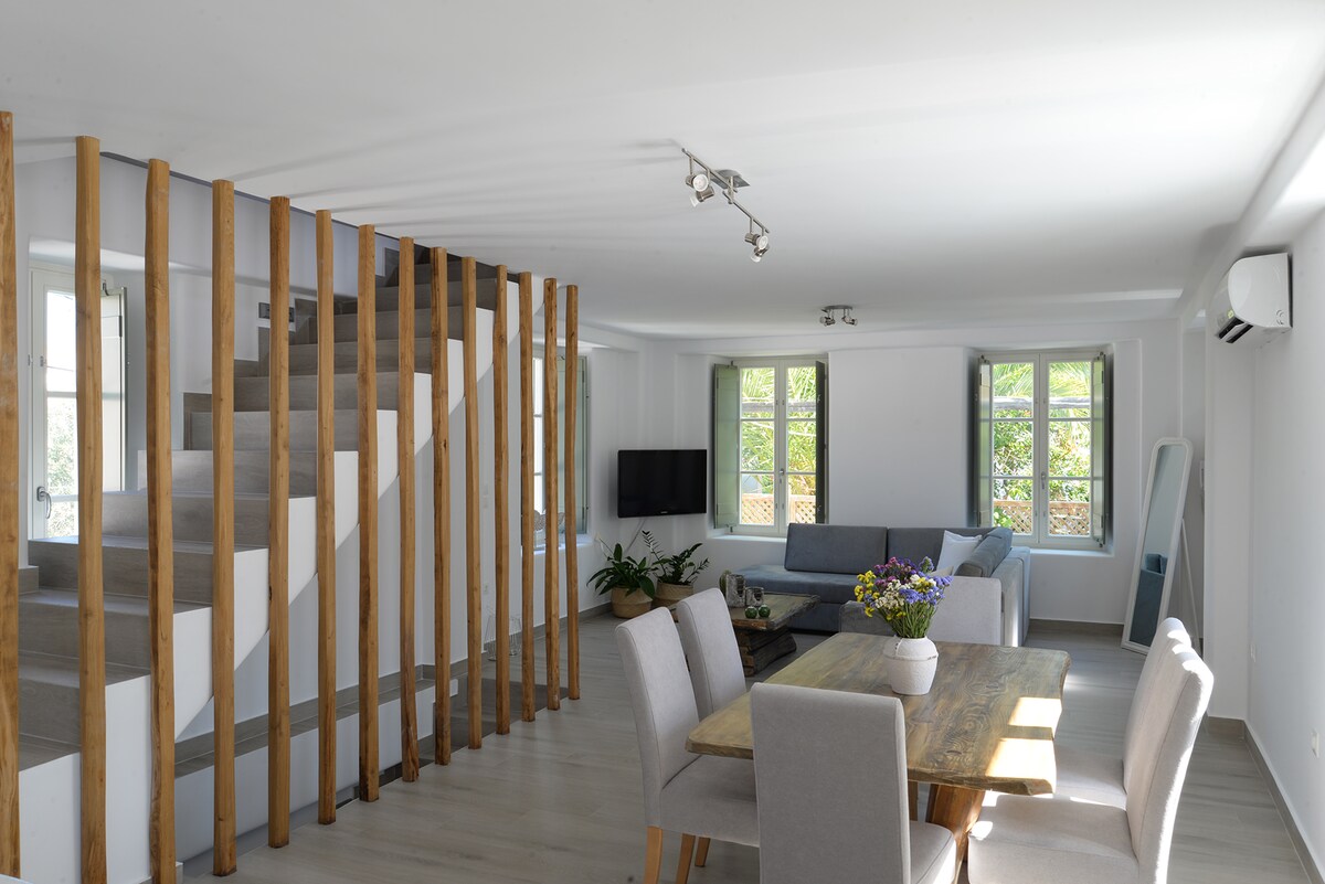 An open living area features a wooden staircase leading to the upper level. Natural light fills the space through windows, illuminating a comfortable seating arrangement and a dining table adorned with a vibrant flower centerpiece.