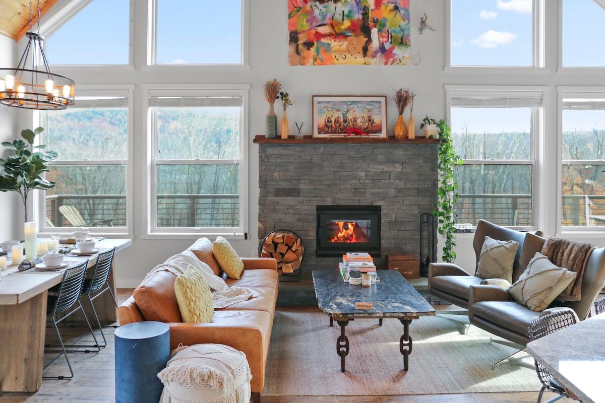 An inviting living area features a stone fireplace, flanked by large windows that provide scenic views. A combination of leather and fabric seating enhances comfort, while a rustic wooden coffee table sits at the center. Natural light fills the space, creating a warm atmosphere.