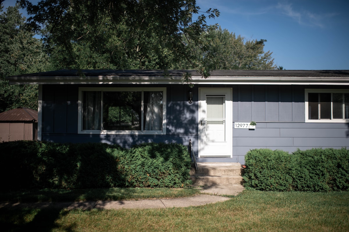 A single-story home is depicted, featuring a light blue exterior and a central white door. A concrete pathway leads to the entrance, bordered by neatly trimmed green bushes. Large windows allow for natural light, and a simple landscaping complements the inviting facade.