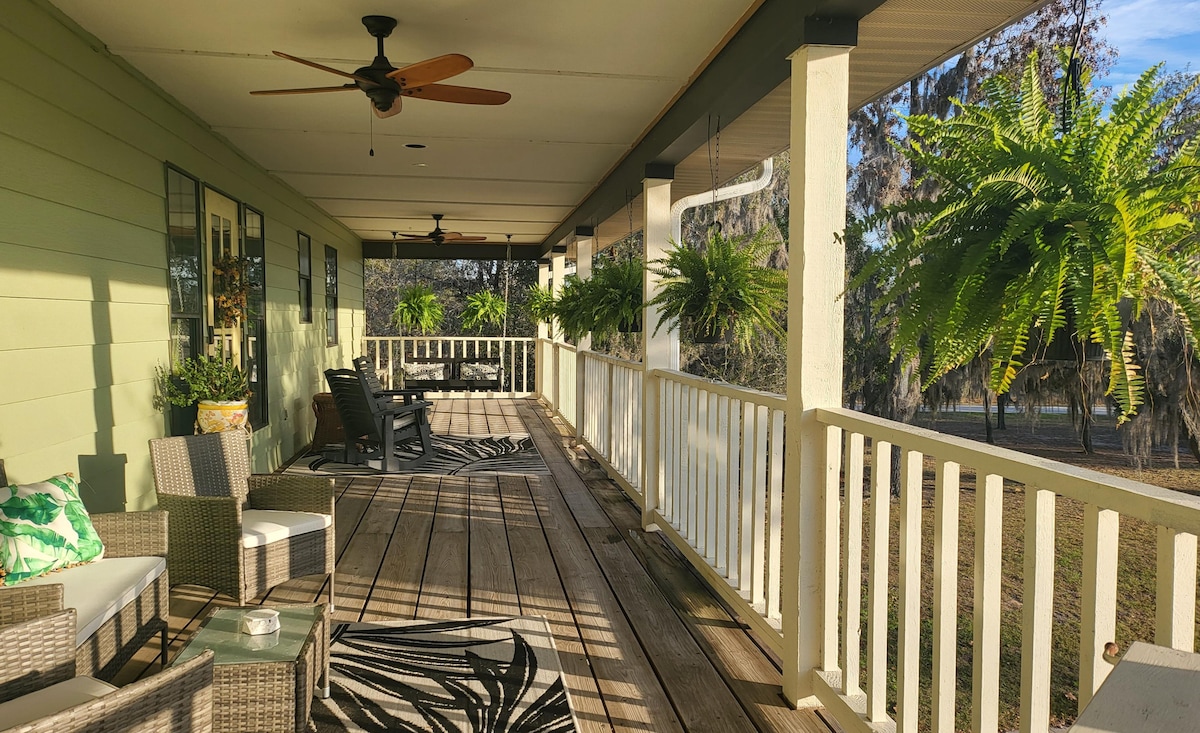 A spacious front porch features comfortable seating arrangements and decorative plants. The wooden decking is enhanced by overhead fans, providing a serene space for relaxation. Lush ferns hang from the railing, and the surrounding natural view contributes to a sense of openness.