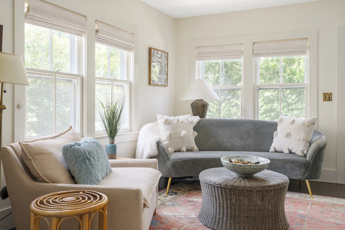 A bright living area is showcased with a light-colored couch and a curved gray sofa, both adorned with decorative pillows. Natural light filters through two windows, highlighting a round woven coffee table and a decorative bowl at its center.