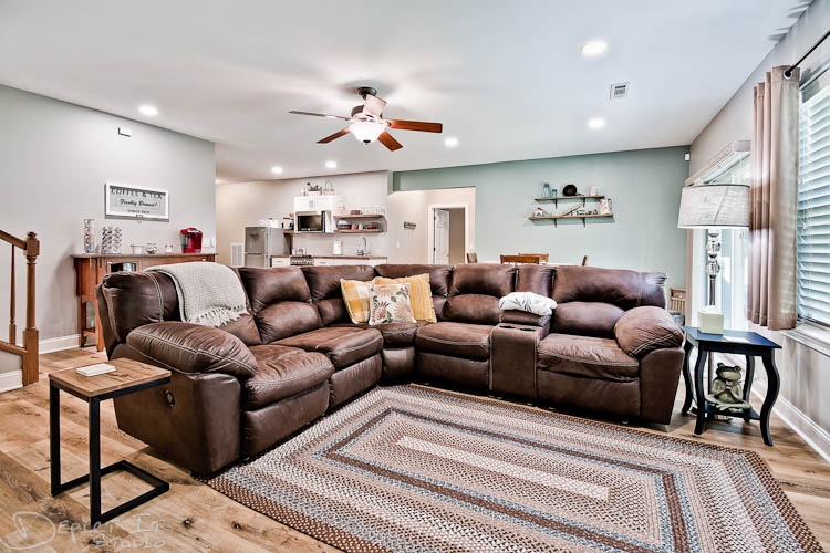A spacious living room features a large, brown sectional sofa arranged around a cozy area rug. A ceiling fan provides airflow, while a light green wall complements the room. A small side table stands nearby, and a doorway leads to an open kitchen and dining area.
