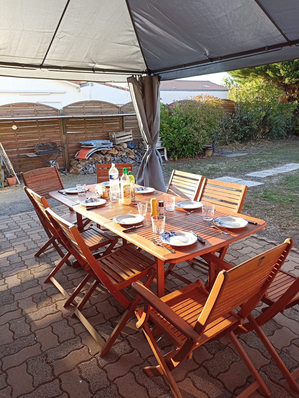 An outdoor dining area features a wooden table set with plates and glasses. Six matching wooden chairs are positioned around the table under a canopy, casting soft shadows on the patio flooring. The garden backdrop includes greenery and storage, enhancing the serene environment.