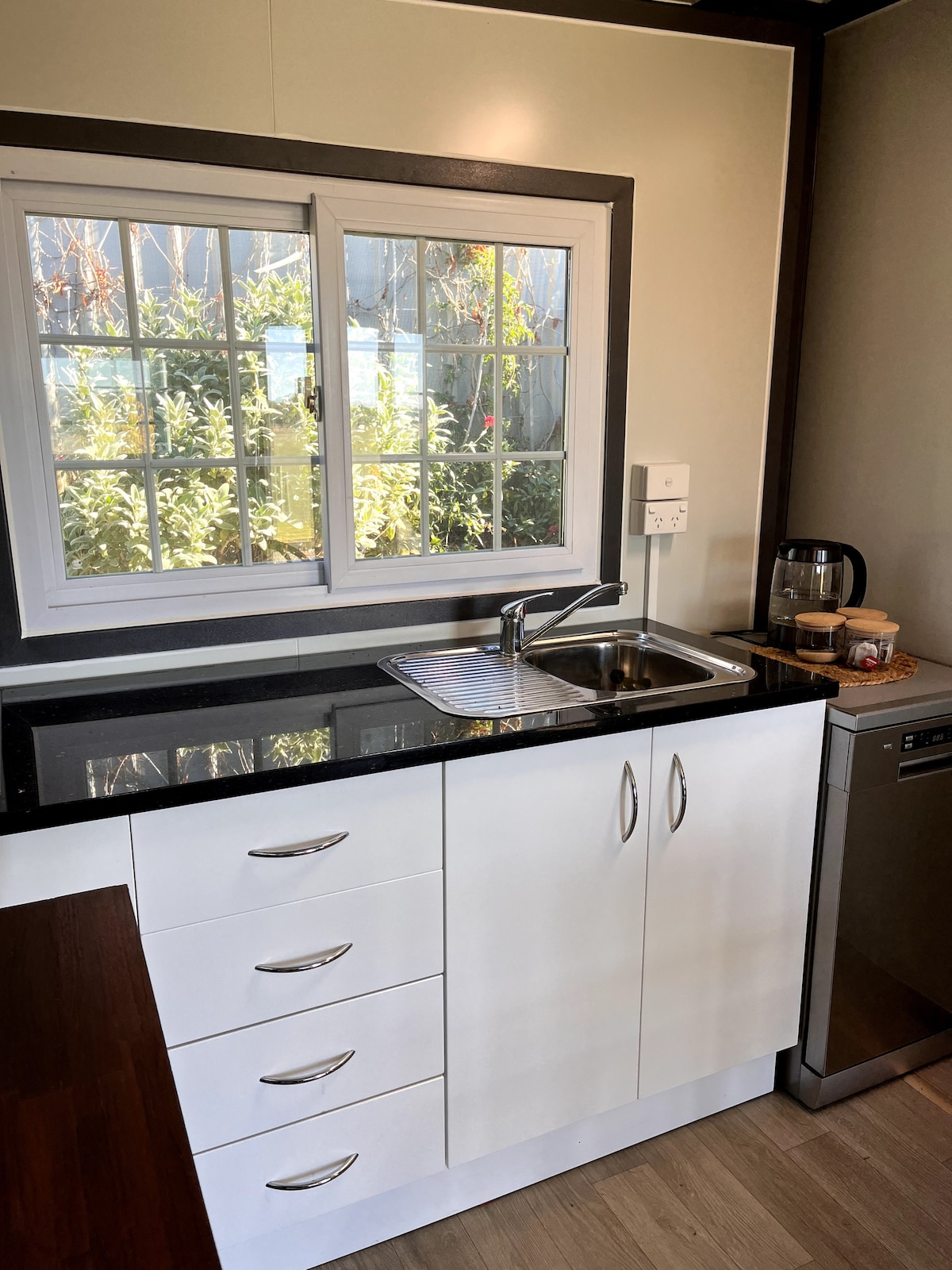 A compact kitchen area features modern cabinetry with a stylish black countertop. A window allows natural light to illuminate the space while offering a view of greenery outside. Stainless steel appliances, including a fridge and a kettle, are positioned neatly alongside the sink.