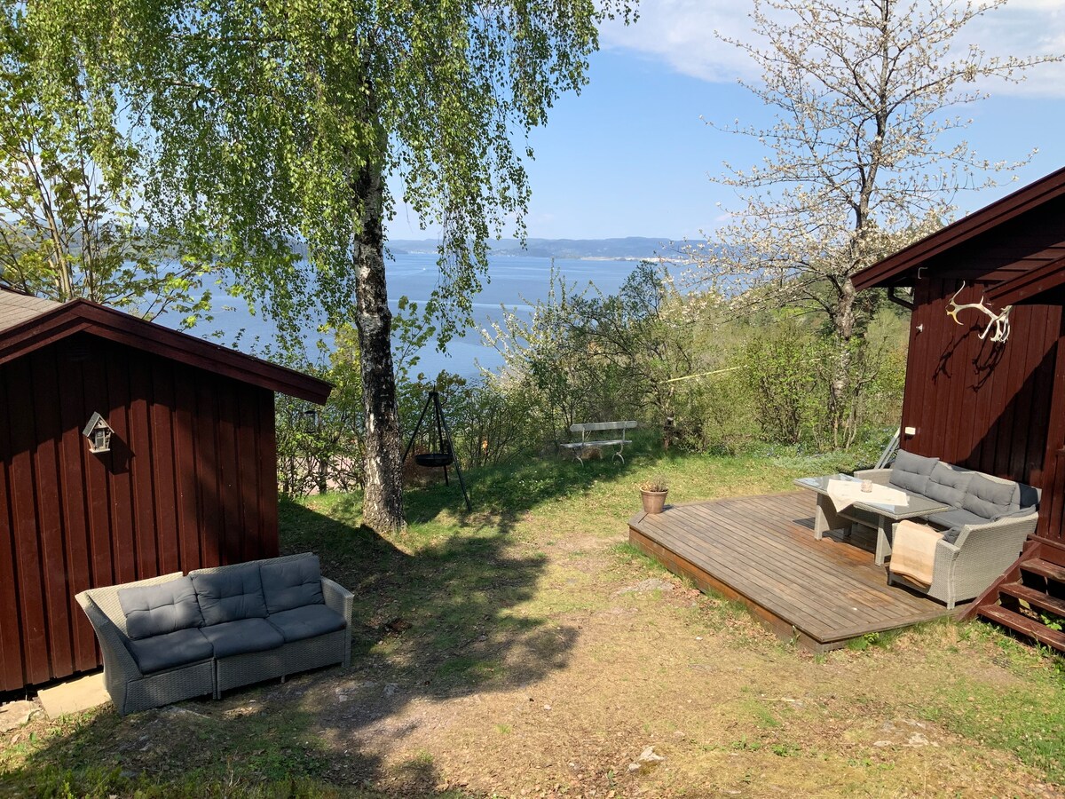 A grassy outdoor area features comfortable seating arrangements, including a sectional sofa and a wooden deck with a table. Surrounding trees provide shade, while the view opens to a serene body of water, contributing to a peaceful outdoor setting.