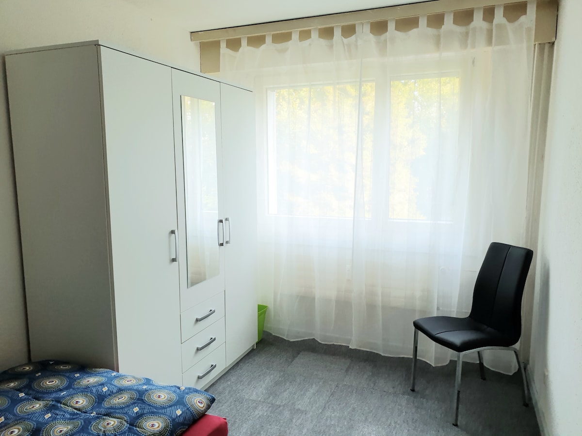 A bright room is equipped with a spacious bed covered in a patterned blanket. A sleek wardrobe with mirrored doors stands nearby, and a black chair is positioned next to a window draped with sheer curtains, allowing natural light to fill the space.