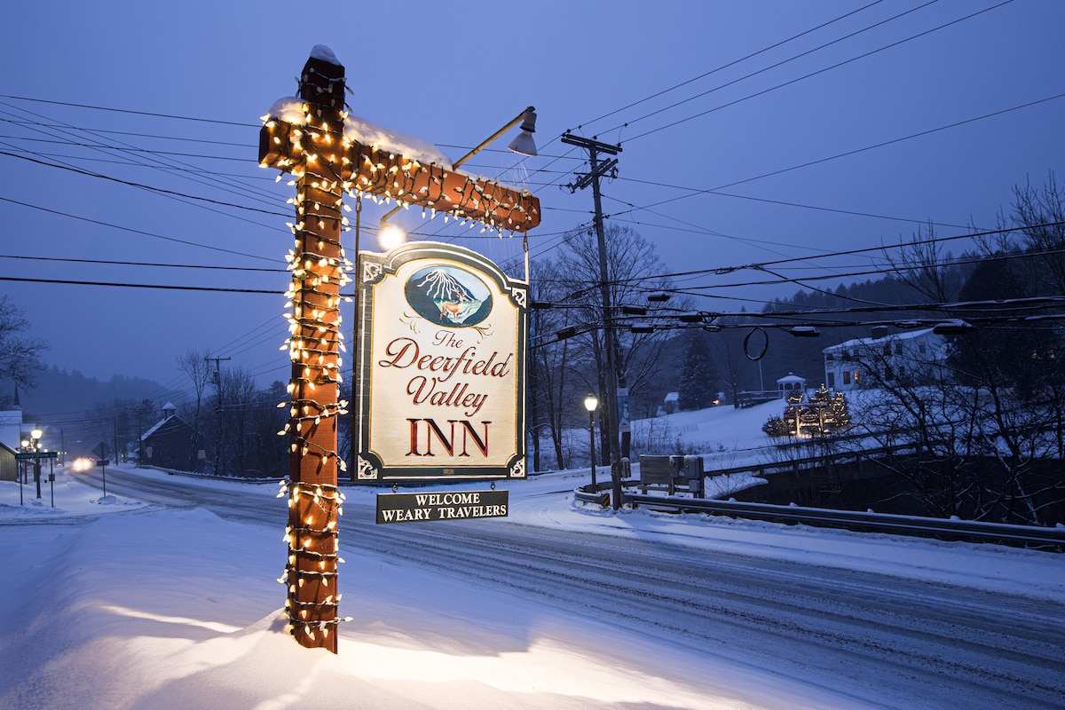 A warmly lit sign for the Deerfield Valley Inn is displayed along a snowy roadside, welcoming travelers. Soft snow blankets the ground and tree branches, while surrounding structures are subtly illuminated in the evening light, creating a serene winter scene.