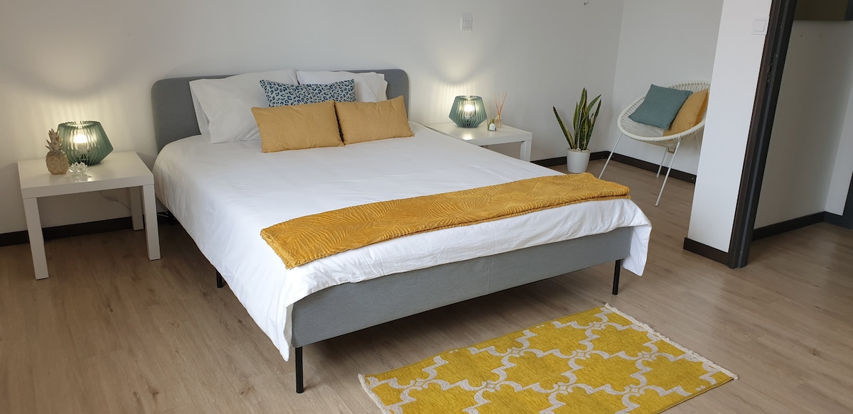 A spacious double bedroom is presented with a queen size bed featuring a white linen sheet and mustard accent pillows. Two bedside tables are positioned on either side, complemented by softly glowing lamps. A patterned yellow rug adds warmth, while potted plants enhance the calm atmosphere.