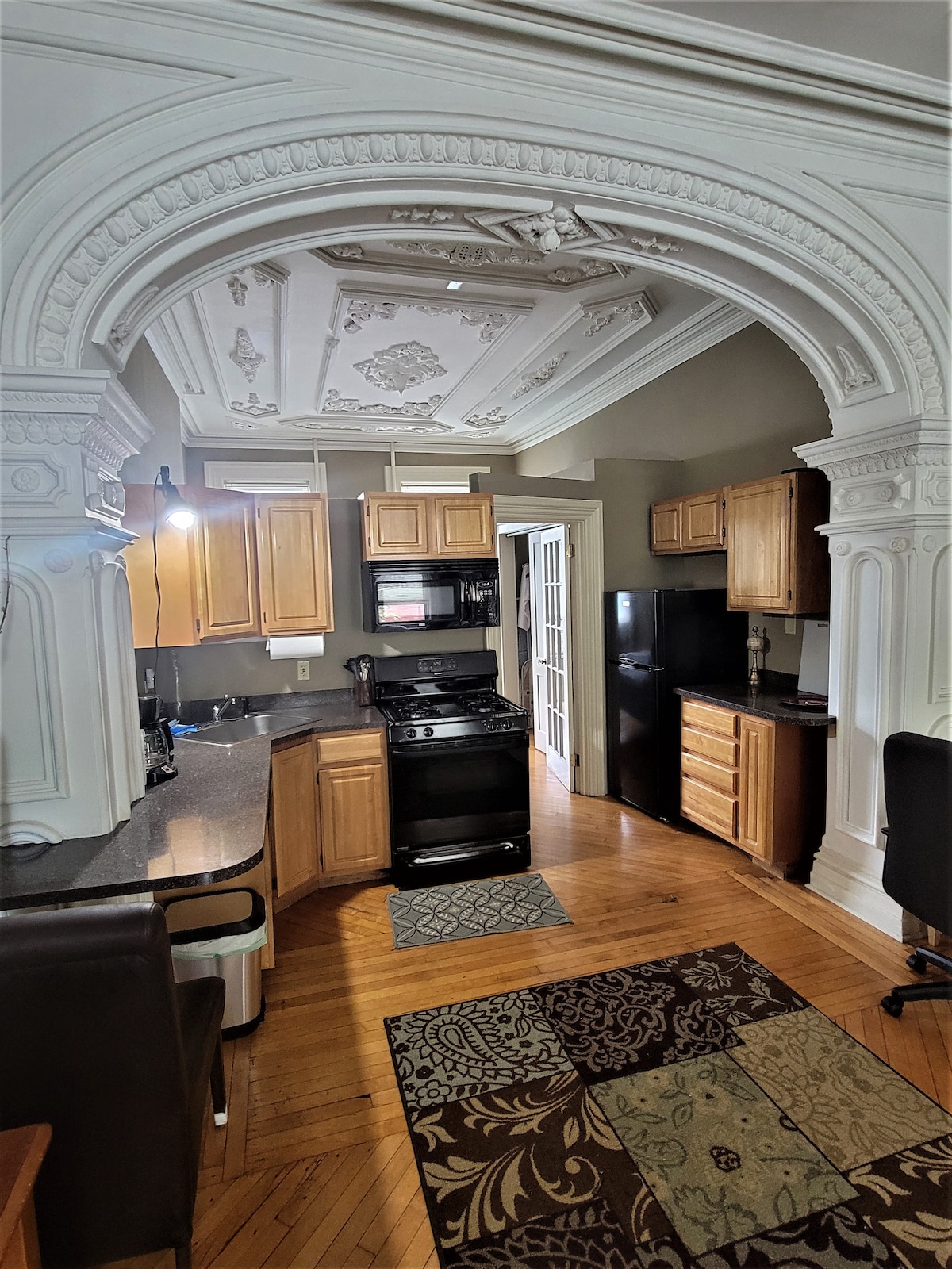 A newly remodeled kitchen features wooden cabinetry and modern appliances. An ornate ceiling adds character above, while a patterned area rug lies on the polished hardwood floor. A dining area is visible, with a dark wood table and chairs positioned nearby.