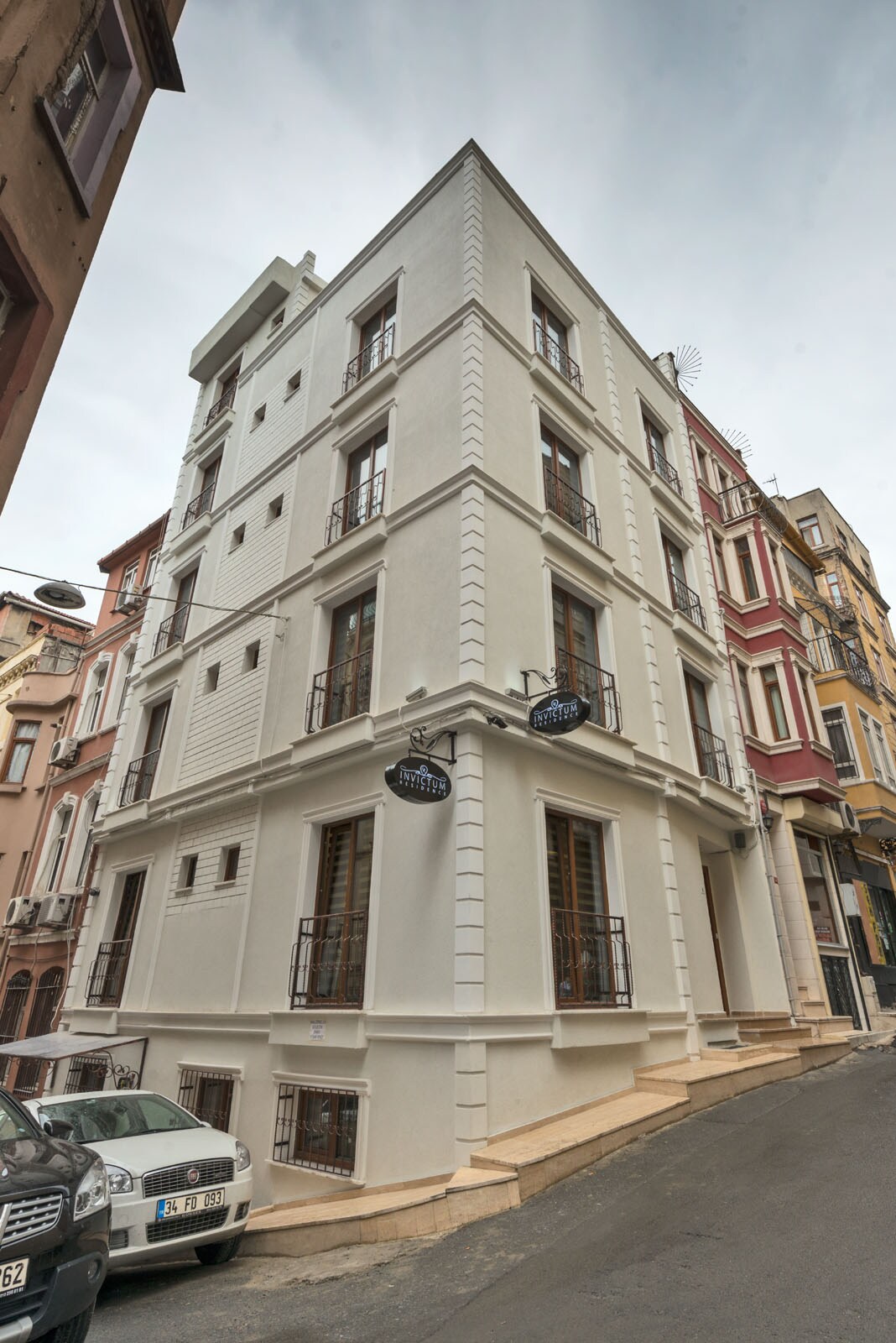 The exterior of a modern building is showcased, featuring a clean white facade with multiple windows and a symmetrical design. The structure has a slightly angled view, and balconies are visible on several floors. A narrow street runs alongside, lined with parked cars.
