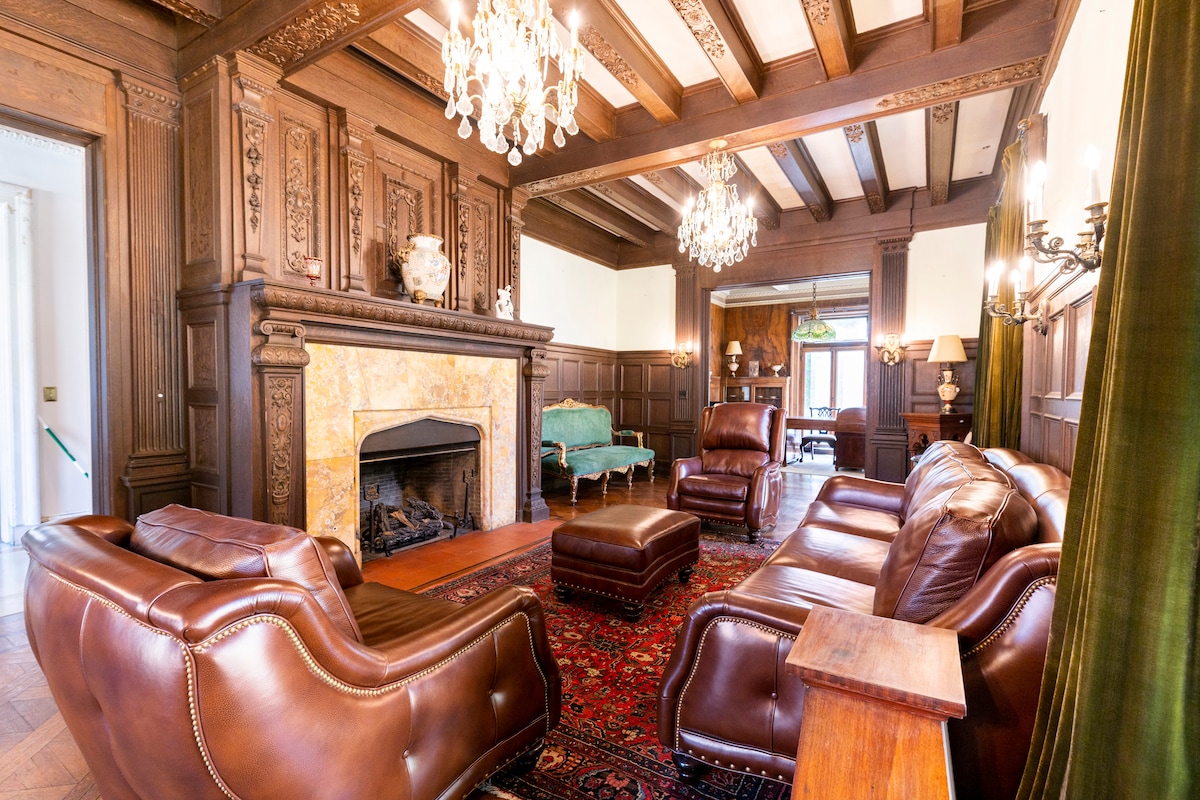A spacious living room exhibits rich wooden paneling and ornate ceiling beams. Plush leather armchairs and a large ottoman are arranged around a decorative fireplace. Two ornate chandeliers hang above, and a vibrant area rug adds color to the hardwood floor.