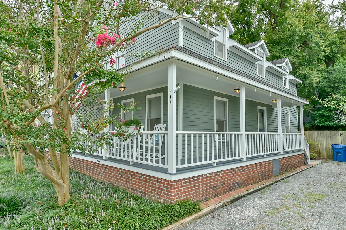 A charming duplex features a wraparound front porch with rocking chairs, surrounded by well-kept greenery and blooming flowers. The exterior showcases a light grey facade with white trim, providing a cozy and inviting presence on a quiet street.