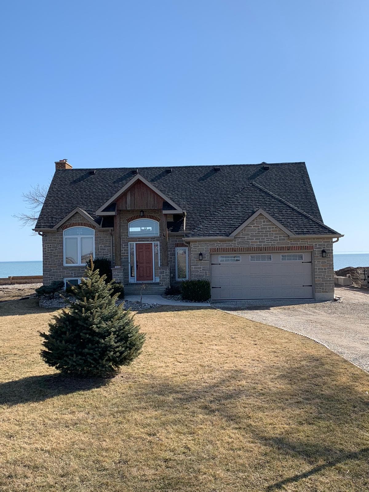 A modern lake house is depicted with a stone exterior and a welcoming entryway. The front yard features a small evergreen tree, while the expansive view of Lake Erie is visible in the background, showcasing an inviting outdoor space.
