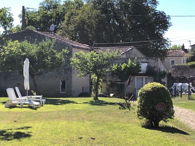 The image showcases the tranquil garden area of the property, featuring a manicured lawn with lounge chairs positioned under sunlight. A hedge adds a natural border, while trees provide shade in the backdrop. The stone buildings of the lodge are visible, adding to the rustic charm.