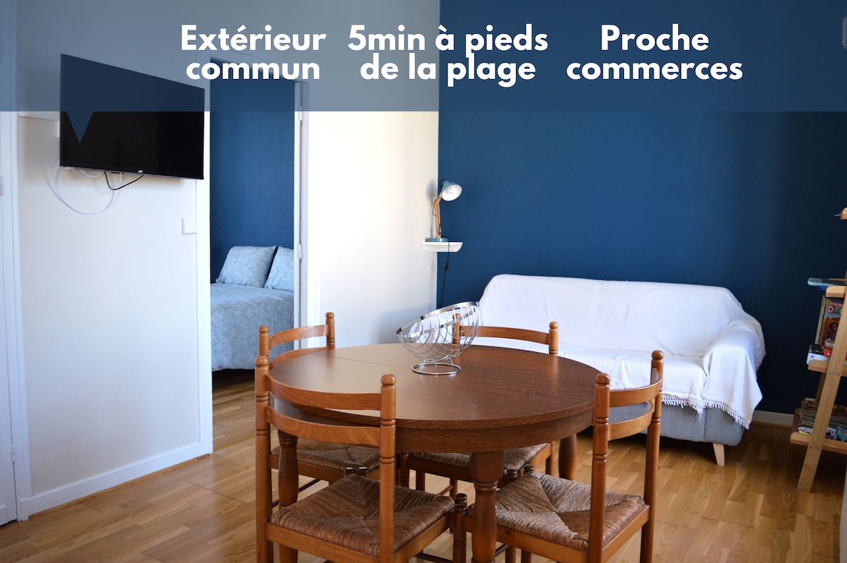 The image displays the living area of the apartment, featuring a round wooden dining table with four chairs. A white sofa is present in the background, and a blue accent wall creates a soothing atmosphere. A wall-mounted television is also visible.