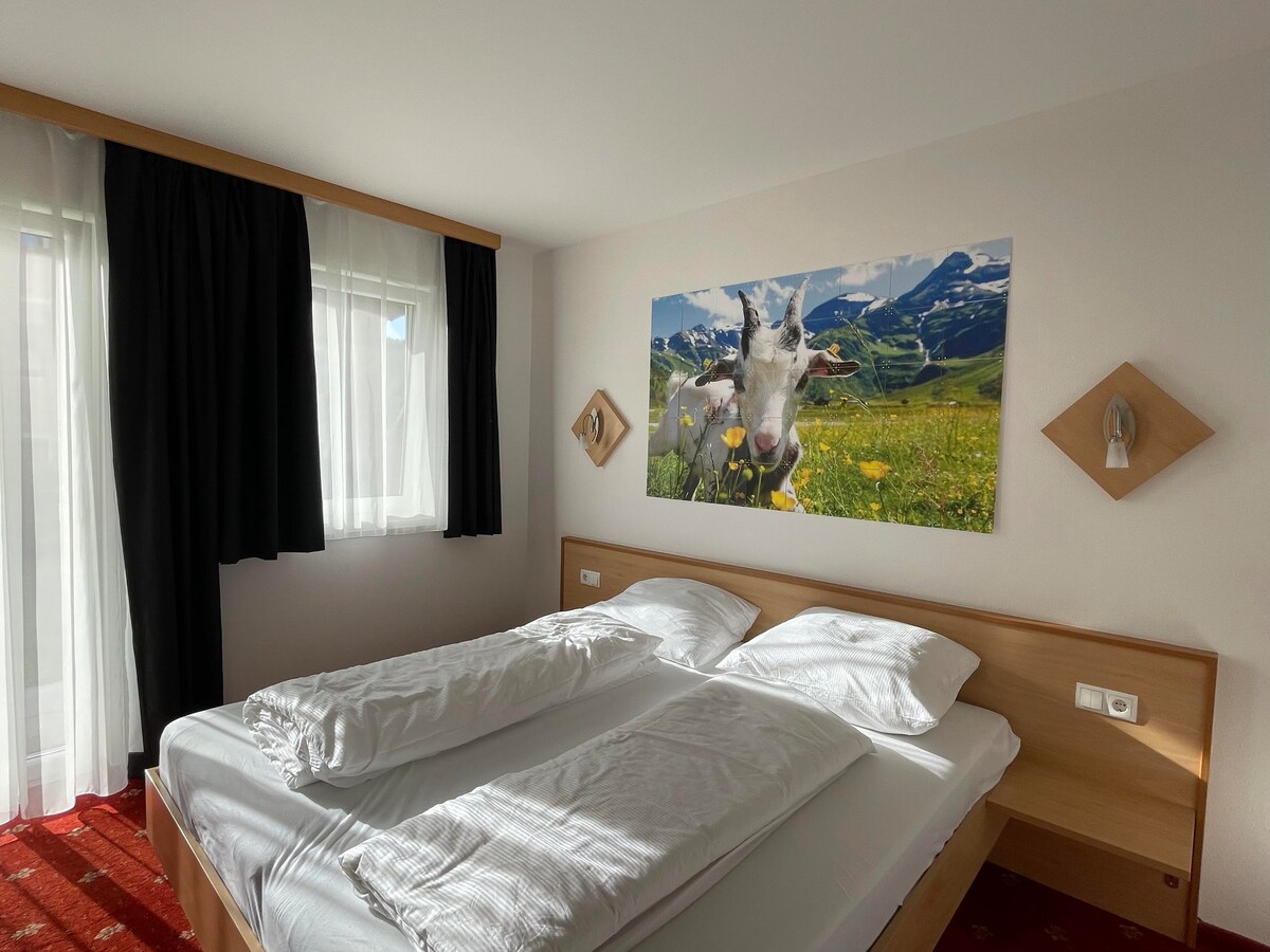 A bright bedroom is featured, showcasing two spacious beds with crisp white linens. Light enters through a large window dressed with simple black curtains. A colorful cow-themed artwork decorates the wall above the bed, contributing to the room’s charm.