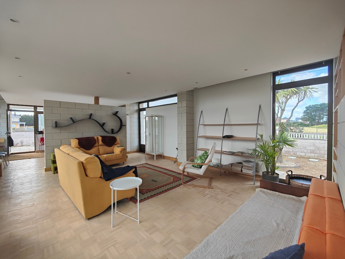 A modern living space is presented with large windows allowing natural light to fill the room. A bright orange sofa is positioned next to a light wooden coffee table. A small plant decorates the side, while a minimalist shelving unit is displayed against the wall.