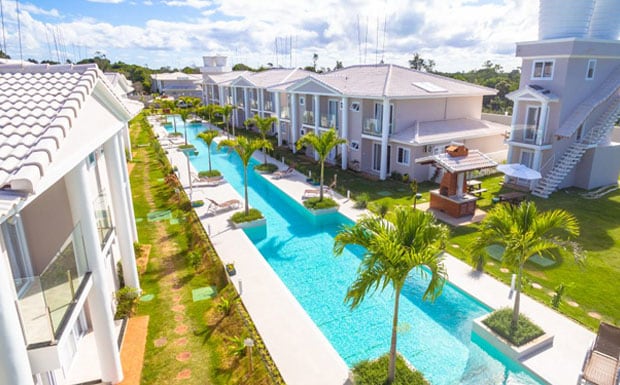 An aerial view captures a neatly arranged complex featuring a long, shimmering pool lined with palm trees. Spacious units are visible on either side, with lounge chairs placed strategically along the pool's edge. A barbecue area is situated in the communal outdoor space.