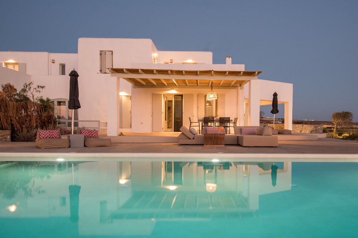 A spacious outdoor patio is visible, featuring comfortable seating arrangements and a dining area beneath a pergola. The serene pool reflects the evening sky, while the villa’s white exterior creates a modern aesthetic, complemented by soft landscape lighting and nearby lush greenery.