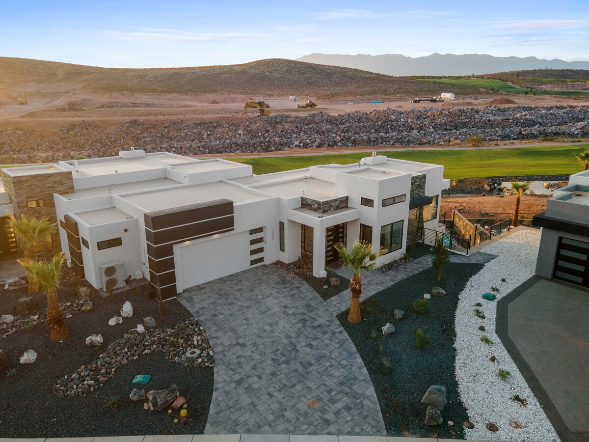 The modern townhome is showcased from an aerial perspective, featuring a spacious driveway and unique architectural lines. Surrounding palm trees enhance the landscape, while the nearby golf course is visible in the background. Pebbled walkways lead to the entrance, creating a distinct exterior design.
