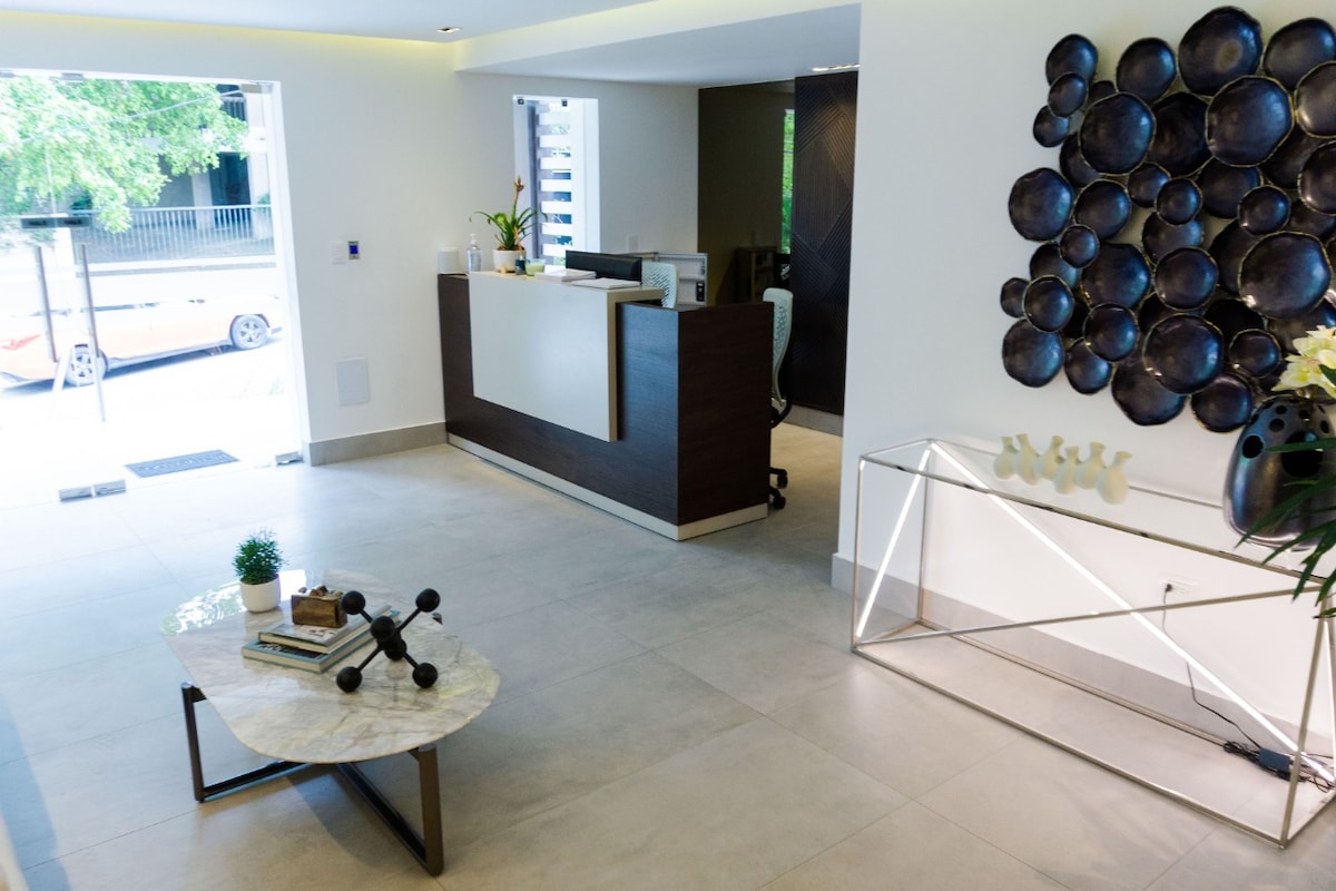 The lobby area is presented with a modern reception desk and a stylish coffee table featuring a marble surface. Decorative elements, including wall art with circular designs, enhance the welcoming ambiance. Natural light floods through the glass entrance, creating a bright and inviting space.