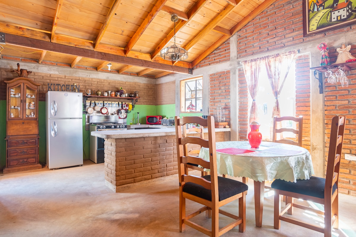 A spacious kitchen featuring high wooden ceilings and exposed brick walls. A dining table with four chairs is positioned near a window adorned with light curtains. The kitchen area includes a fridge, stove, and various shelves displaying dishes and kitchenware.