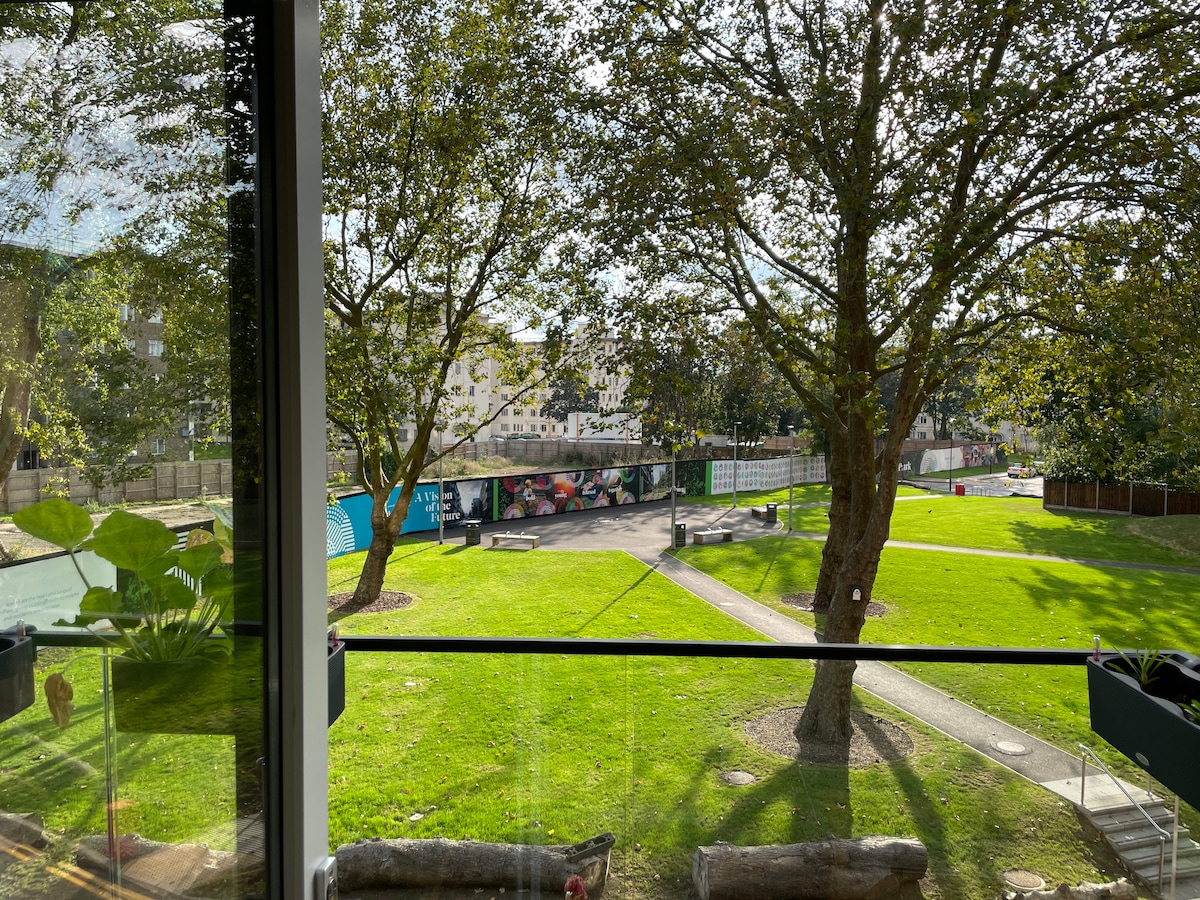 The view from the window showcases a vibrant green park adorned with large trees. A pathway meanders through the grass, leading to colorful murals on display in the background. Natural light floods the scene, creating a serene outdoor atmosphere.