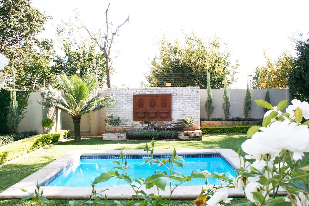 A well-maintained garden features a swimming pool surrounded by lush greenery and decorative plants. A stone wall with three carved faces adds an artistic focal point, while white roses gently frame the view, enhancing the serene outdoor space.