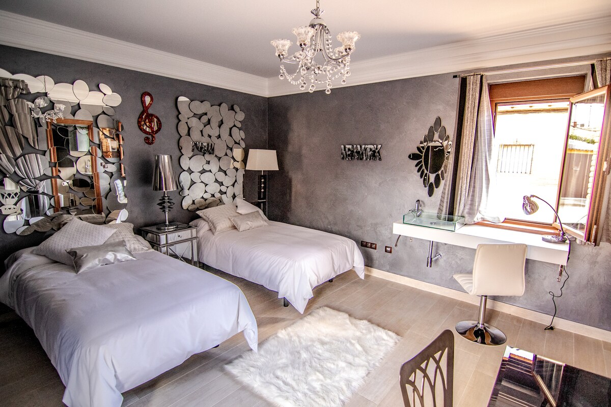 The room features two spacious twin beds, located against a stylish grey wall adorned with decorative mirrors. A window, fitted with light curtains, allows natural light to enter. A desk and chair are placed beside the window, enhancing the functionality of the space.