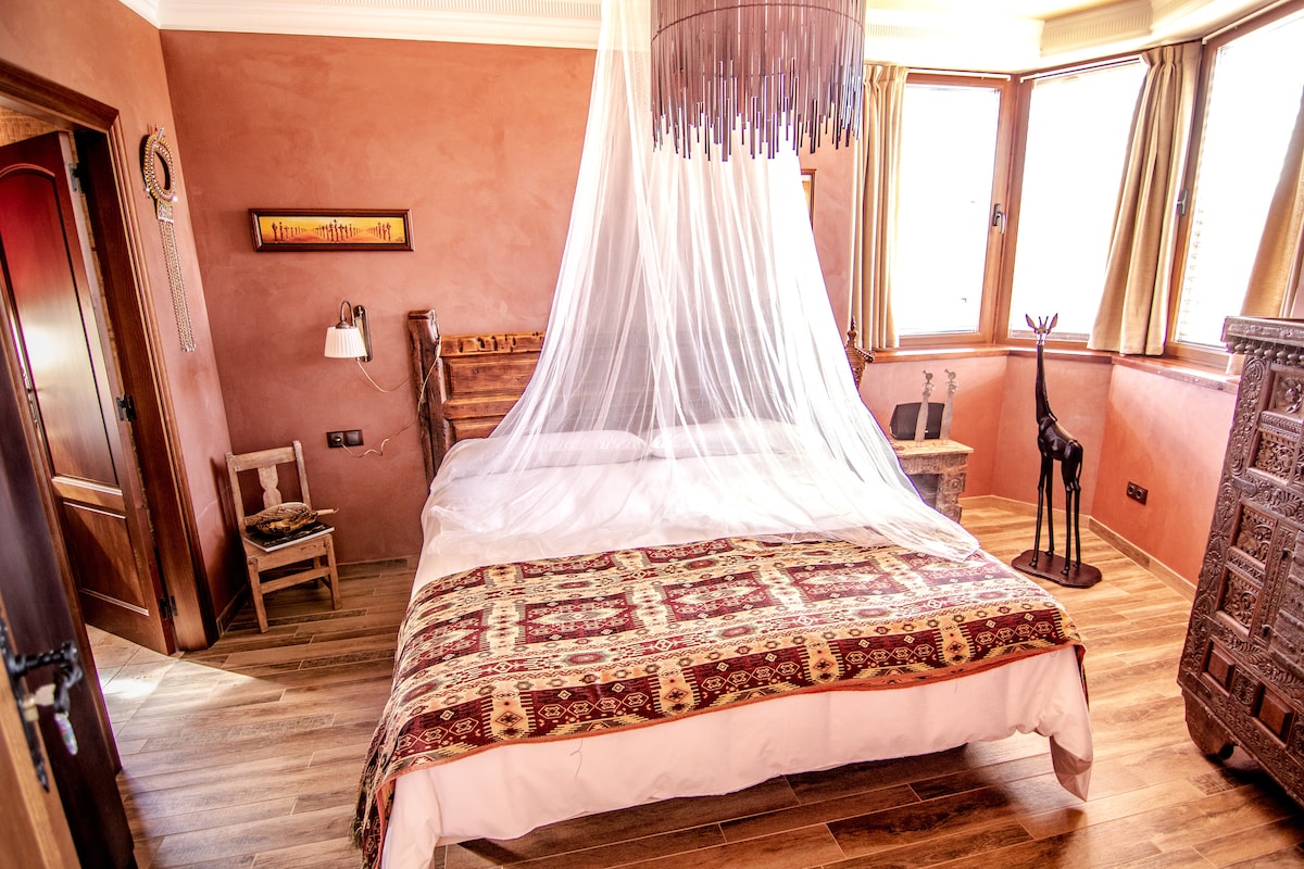 A bedroom featuring a spacious bed draped with a light canopy. Earth-toned walls and wooden furnishings create a calming environment. A colorful patterned bedspread adds visual interest, while large windows allow natural light to enhance the warming atmosphere of the room.