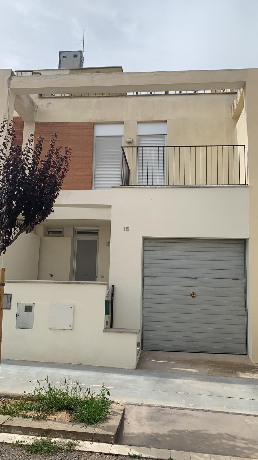 A three-story house features a modern facade with a mix of neutral tones. A garage door is prominently visible, along with a small entrance area. A balcony extends from the upper floor, framed by slender railings, while windows are arranged symmetrically beneath it.