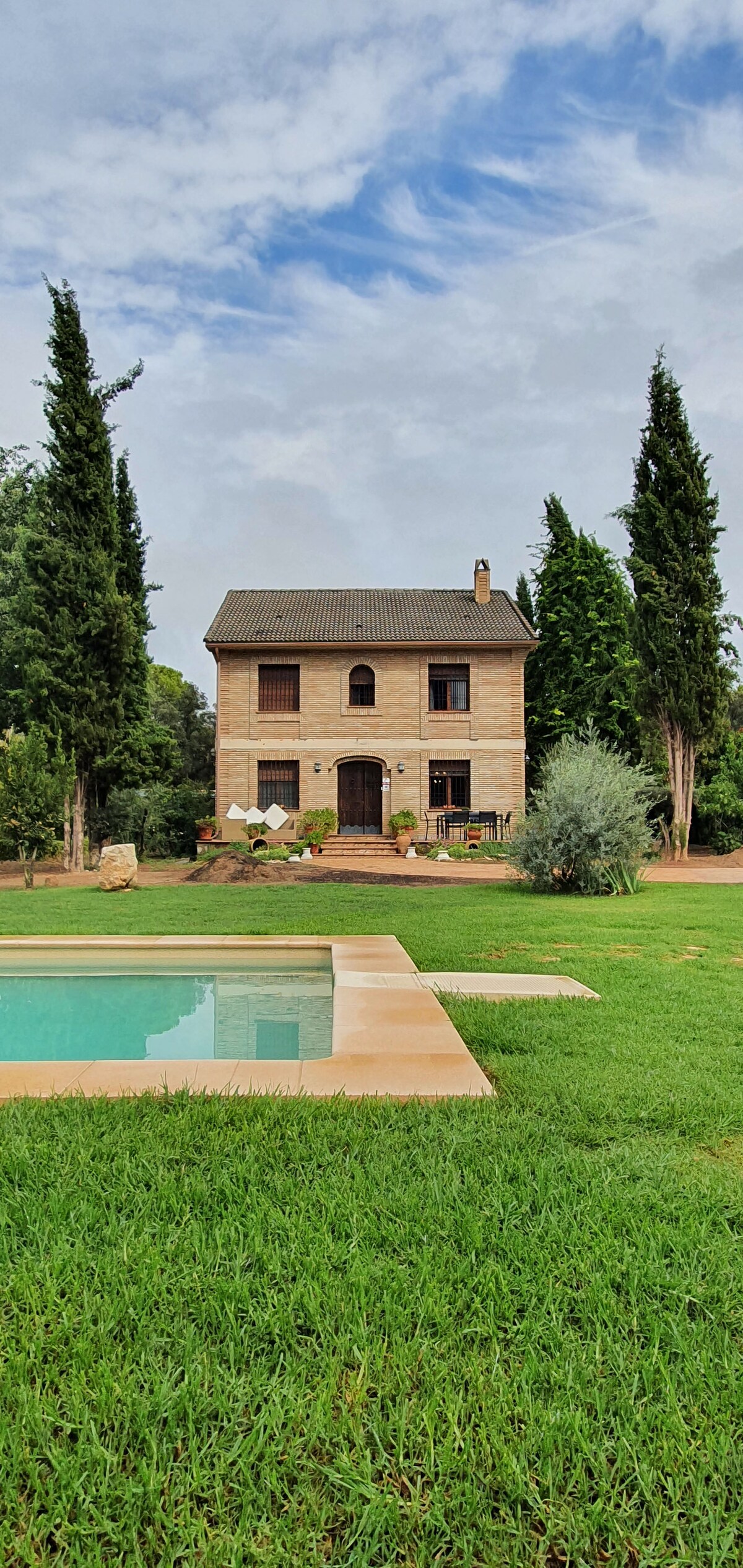 A two-story brick house is surrounded by tall cypress trees, providing a sense of privacy. Lush green grass and a clear pool are visible in the foreground, contributing to a serene outdoor space. The property is designed for relaxation and connection with nature.