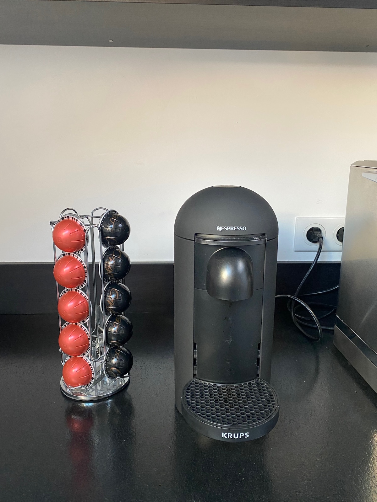 A coffee station is presented with a sleek black Nespresso machine and a metal rack holding vibrant red and black coffee capsules. The dark countertop enhances the contemporary design with electrical outlets visible in the background.