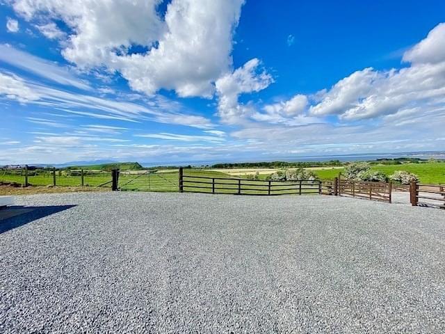 A gravel driveway opens to a spacious area with panoramic views of green fields and distant hills. Blue skies filled with scattered clouds stretch above, enhancing the serene countryside setting.