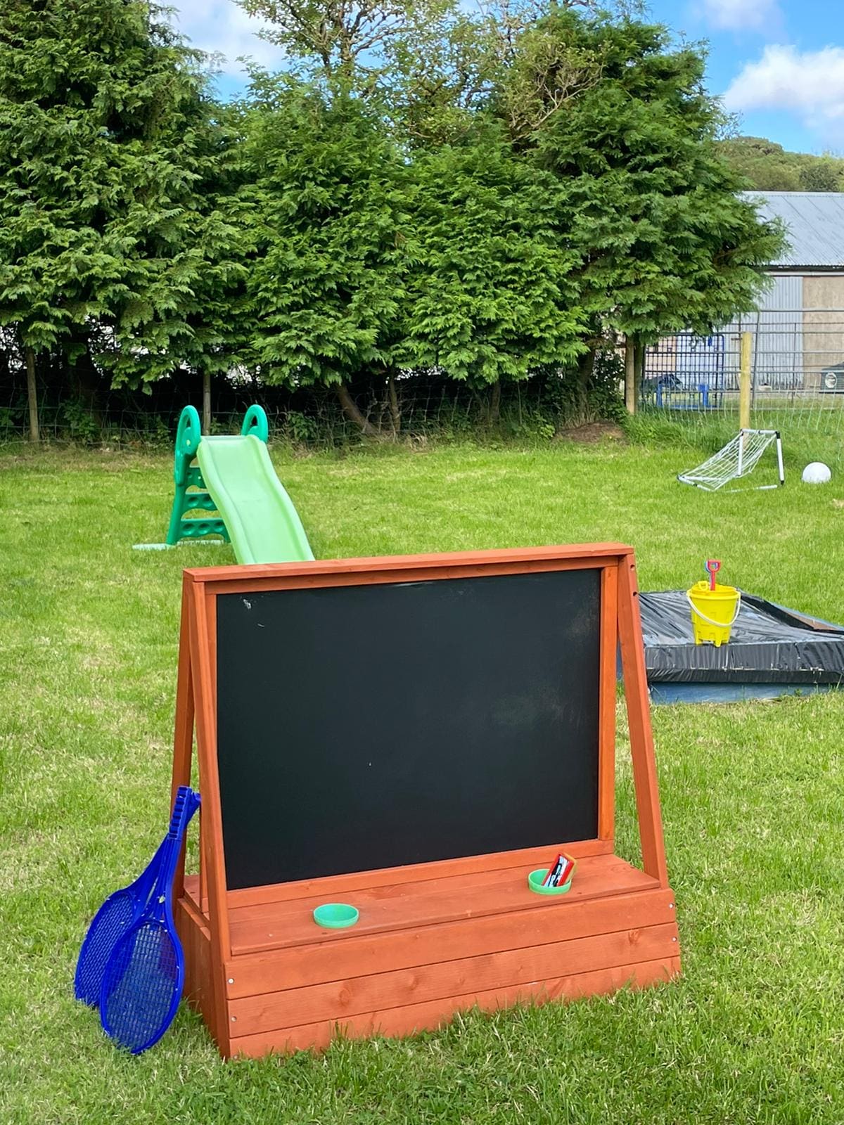 A vibrant outdoor play area is featured, equipped with a green slide, a black chalkboard, and a sandbox. Nearby, a soccer goal is visible, along with various play items, including a blue fishing net. Lush greenery surrounds the space, creating a playful environment for children.