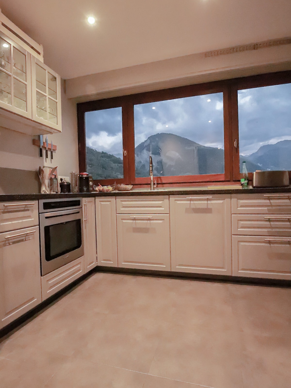 A modern kitchen is showcased with a spacious layout and ample counter space. The cabinetry is light-coloured, complemented by a large window offering panoramic views of the mountains. Appliances such as an oven and stove are integrated, enhancing the kitchen's functionality.