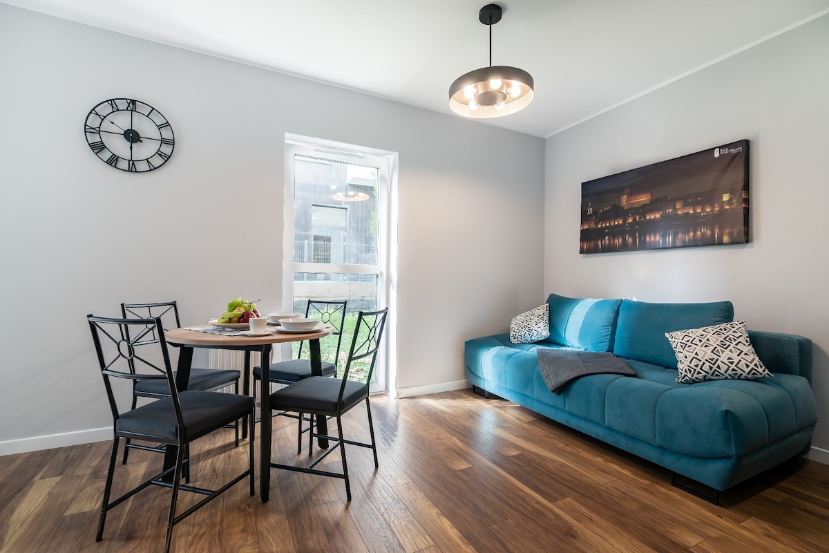 A modern living area is highlighted by a cozy blue sofa beside a round table set for two. A large window allows natural light to brighten the space, while a wall clock adds a functional decor element. Wood flooring contributes to a warm ambiance.