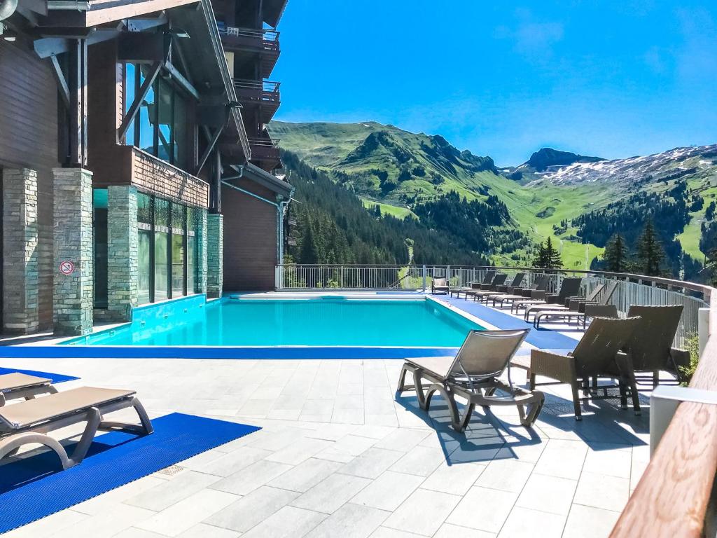 An outdoor pool area is surrounded by mountains and greenery, with lounge chairs positioned along the deck. The clear blue sky reflects off the water, creating a serene atmosphere. Large windows of the building provide a view of the scenic landscape.