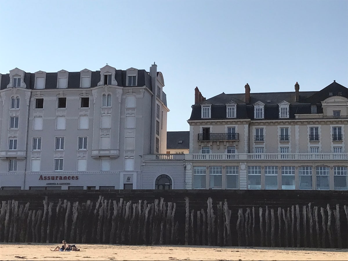 Two buildings are visible along the beach, featuring distinct architectural styles. The first building has a modern façade, while the second showcases classic design elements, including balconies. The sand stretches in the foreground, with beachgoers relaxing at the water's edge.
